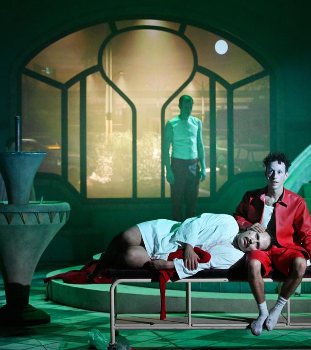 Düstere Theaterszene mit expressionistischem Bühnenbild: Eine Person in einem roten Hemd und roter Hose sitzt barfuß auf einem schlichten Metallbett, mit leerem Blick nach vorne. Eine andere Person in weißem Hemd und dunkler Hose liegt mit dem Kopf auf ihrem Schoß, die Augen geschlossen. Im Hintergrund steht eine weitere Person mit weißem Unterhemd und roter Hose vor einem großen, schlüsselförmigen Fenster, durch das eine nächtliche Stadtszene mit Autos und Straßenlaternen zu sehen ist. Links im Bild ein Brunnen, der Boden ist gefliest. 