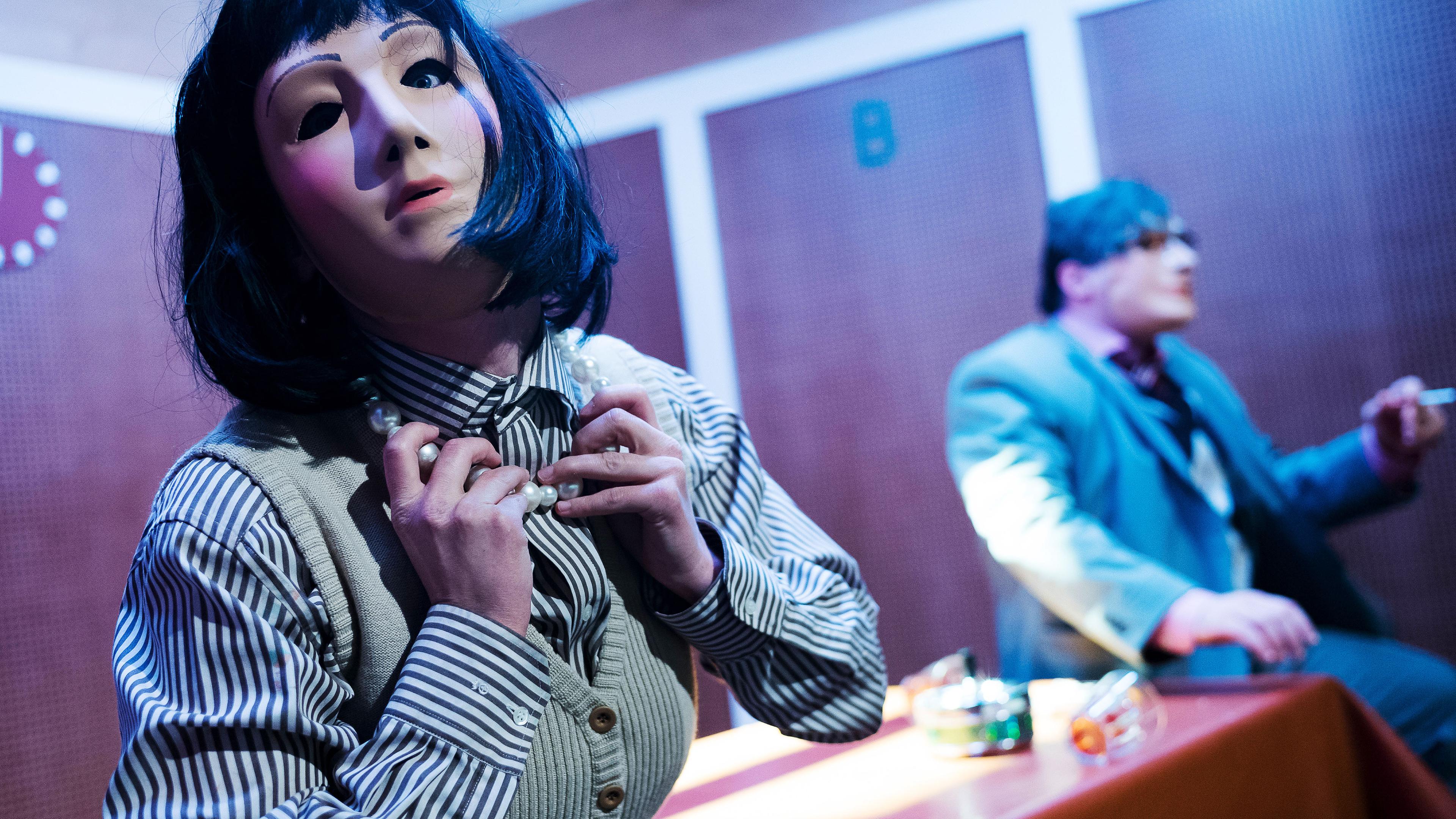 Links im Vordergrund eine Frau mit einer Silikonmaske, die ihr Gesicht vollständig verdeckt. Sie trägt eine schwarze Perücke, einen grau-weiß gestreifte Bluse und eine helle Strickweste. Rechts ein Mann, ebenfalls mit Maske und schwarzer Perücke, er trägt eine Brille und einen grauen Anzug. Zwischen ihnen ein Tisch mit brauner Linoleumtischdecke, darauf zwei umgestürzte Gläser und ein Aschenbecher.  