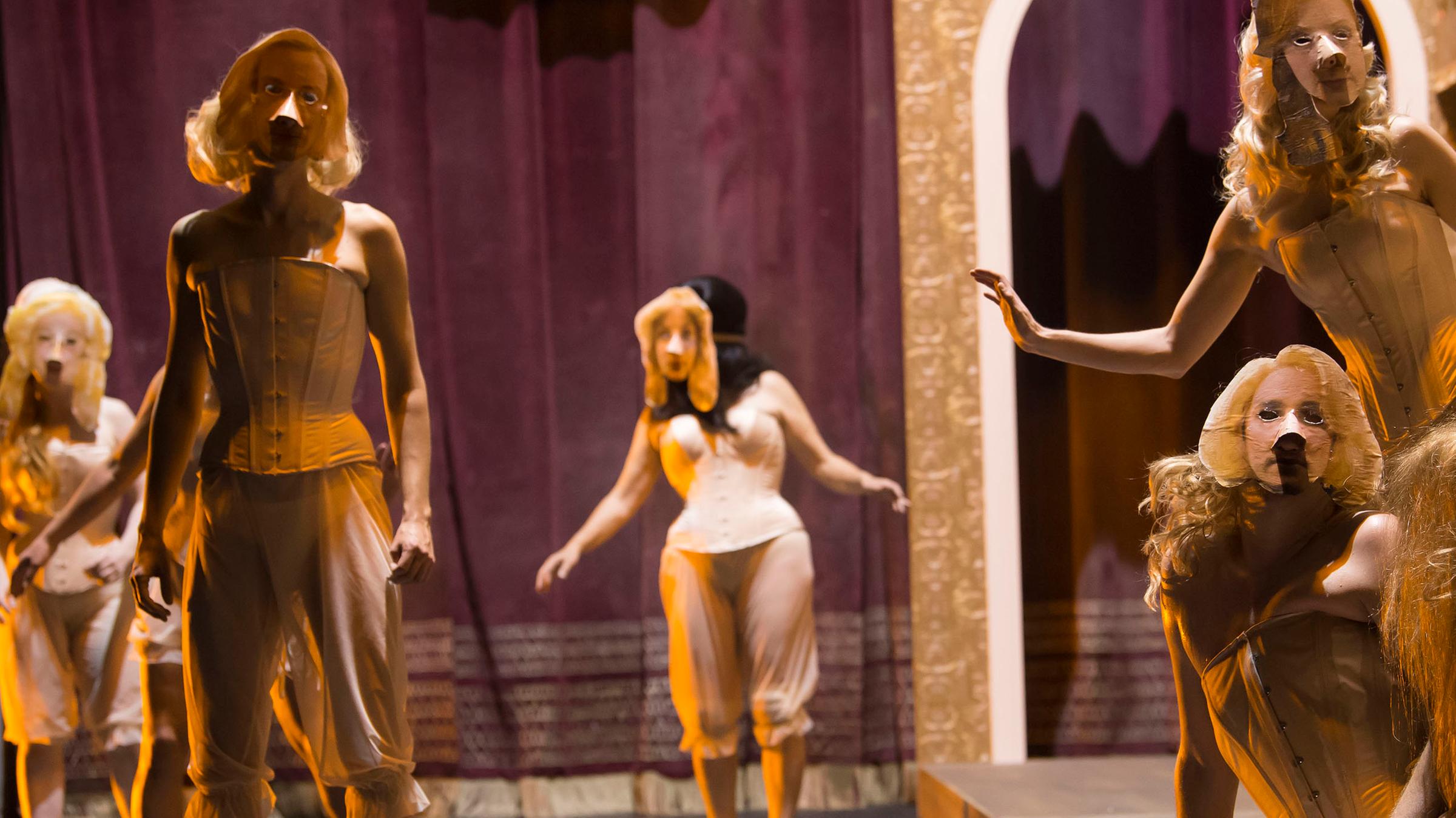 Seitlicher Blick auf die Bühne. Sechs Personen sind dem Betrachter zugewandt , in verschiedeneren Positionen stehend und sitzend. Sie tragen helle Korsagen, Pluderhosen, Plüschpantoffeln sowie Papiermasken.