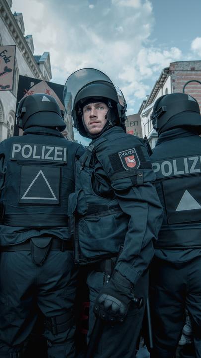 Jan Vogt (Justus Johanssen) steht vor einem mit schwarzer Einsatzkleidung und Helmen bestückten Einsatztrupp der Polizei und blickt nach vorne in die Kamera. Über den Personen sind Demonstrationsschilder und einige Hausfassaden zu erkennen.