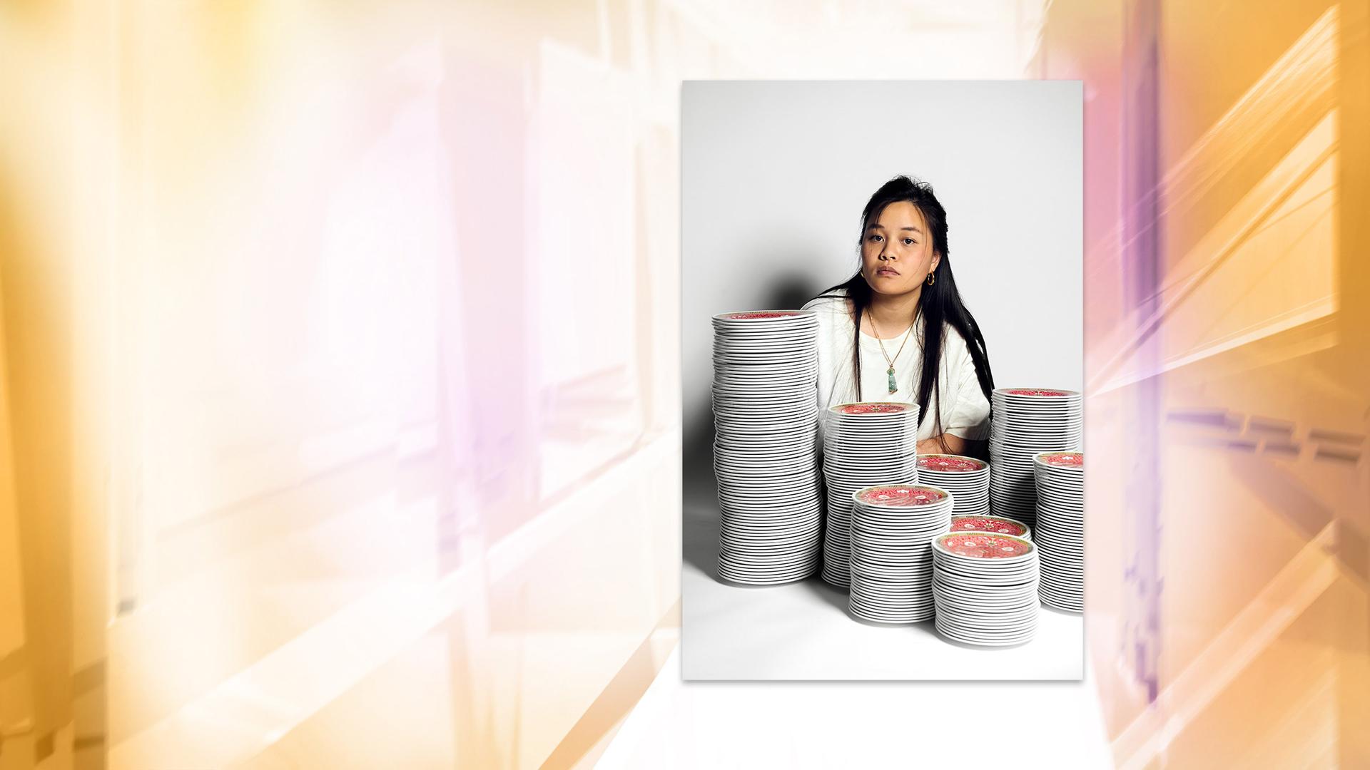Fotografie einer asiatischen Frau mit langen Haaren, die hinter mehreren Stapeln mit bunten Schüsseln sitzt.