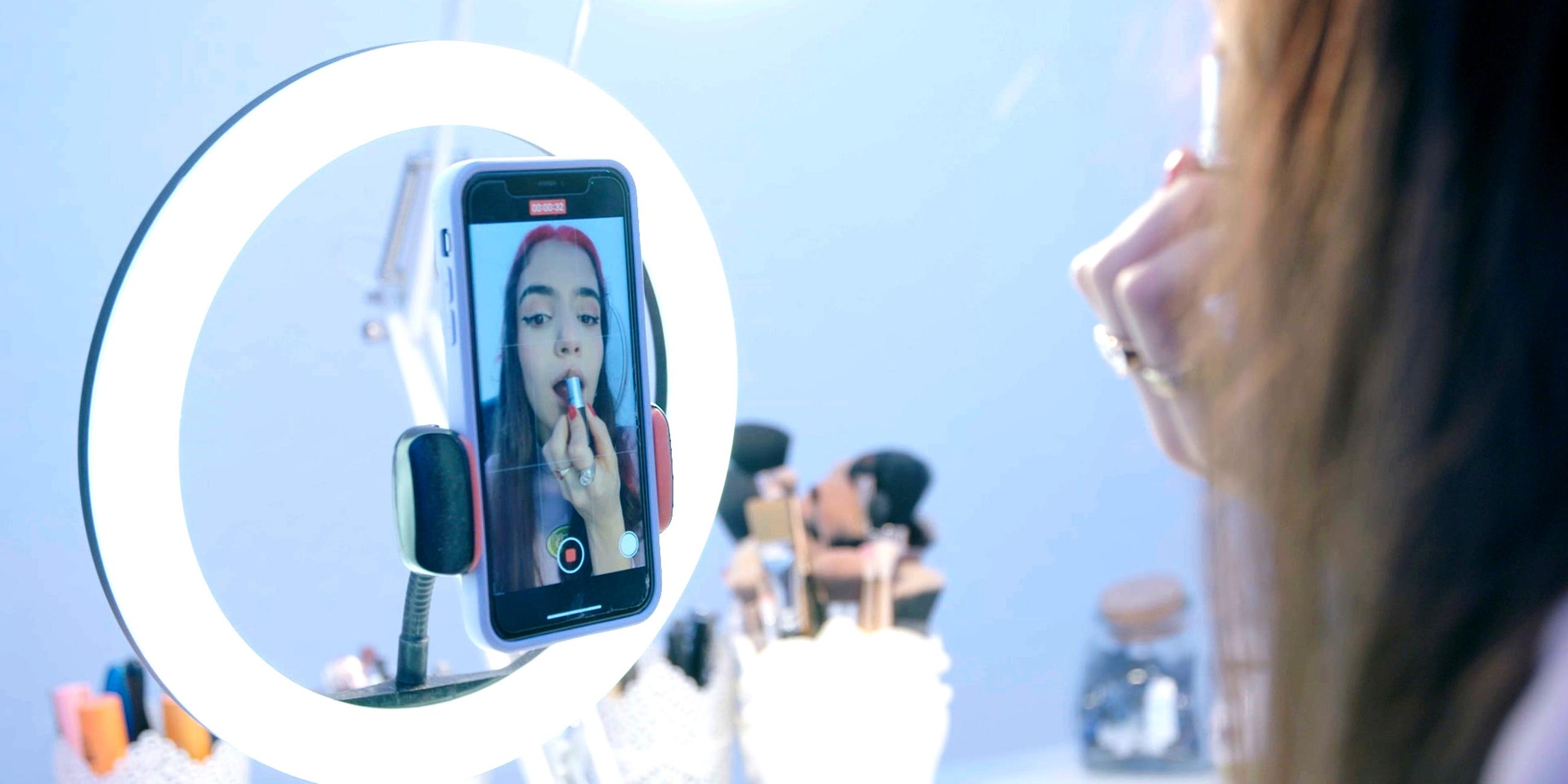  Die 17-jährige Hasti sieht sich in der Innenkamera ihres Handys an und schminkt sich dabei die Lippen. Das Handy ist an einem Ringlicht befestigt und auf einem Tisch befinden sich mehrere Make-up-Pinsel.