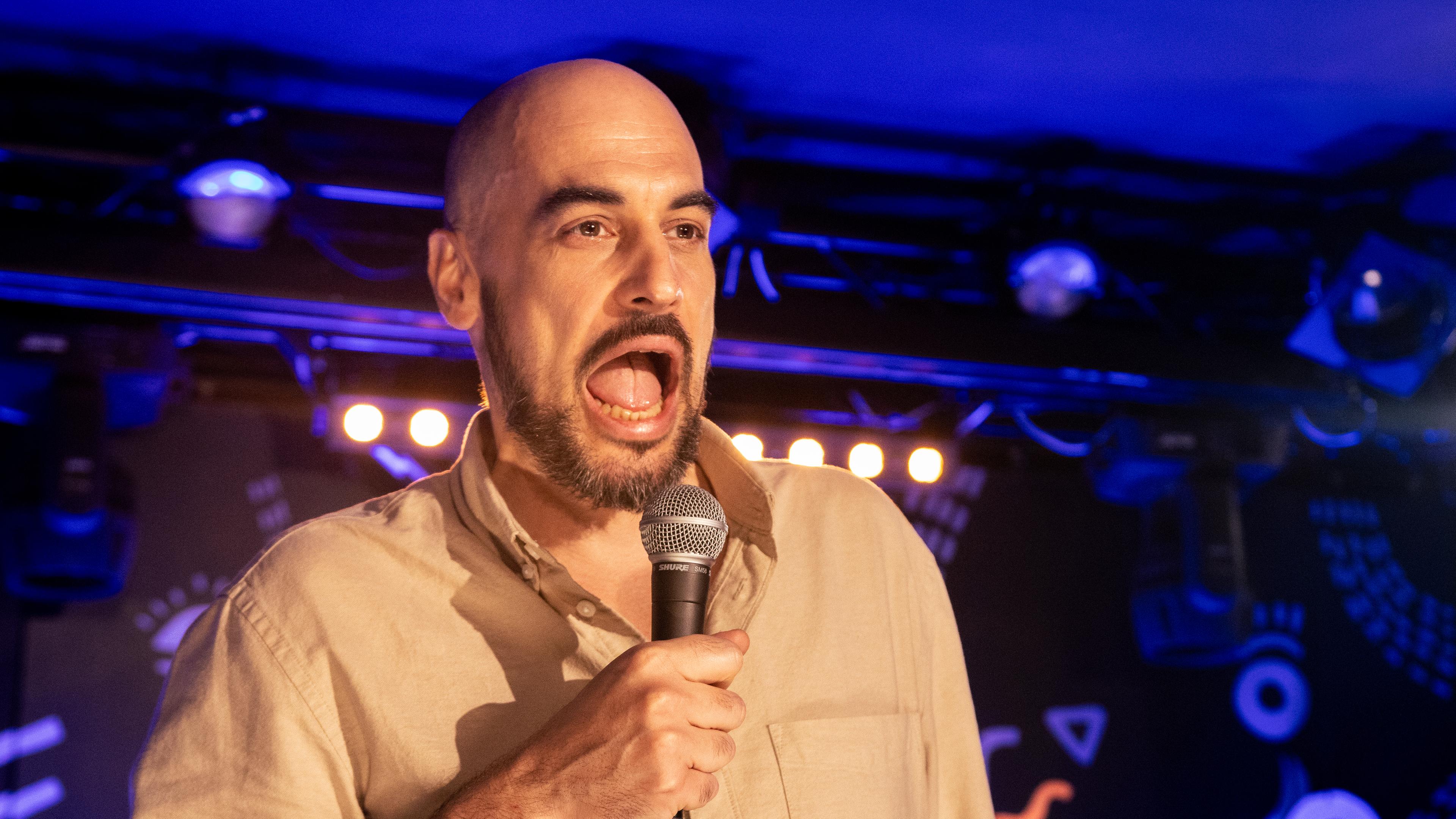 Thomas Kornmaier steht auf der Bühne. Er hat einen offenen Mund und hält in seiner rechten Hand ein Mikrofon.