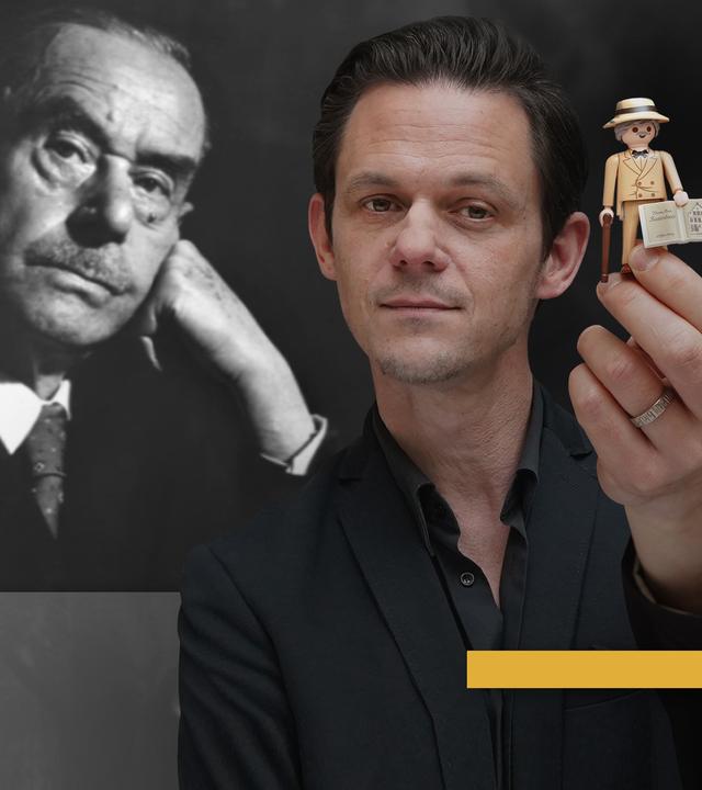 Jo Schück hält eine Playmobil Mann Figur, mit Thomas Mann im Hintergrund