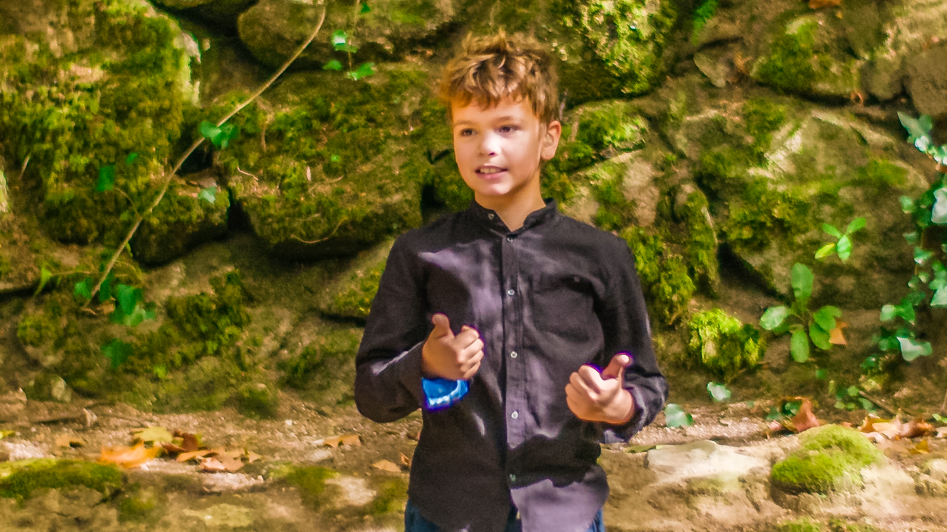 Tiago steht vor einer moosbewachsenen Felswand im Wald. Hier will er seine erste Unterrichtsstunde halten. Er trägt eine Jeans und ein dunkles Hemd und wirkt konzentriert und zuversichtlich.