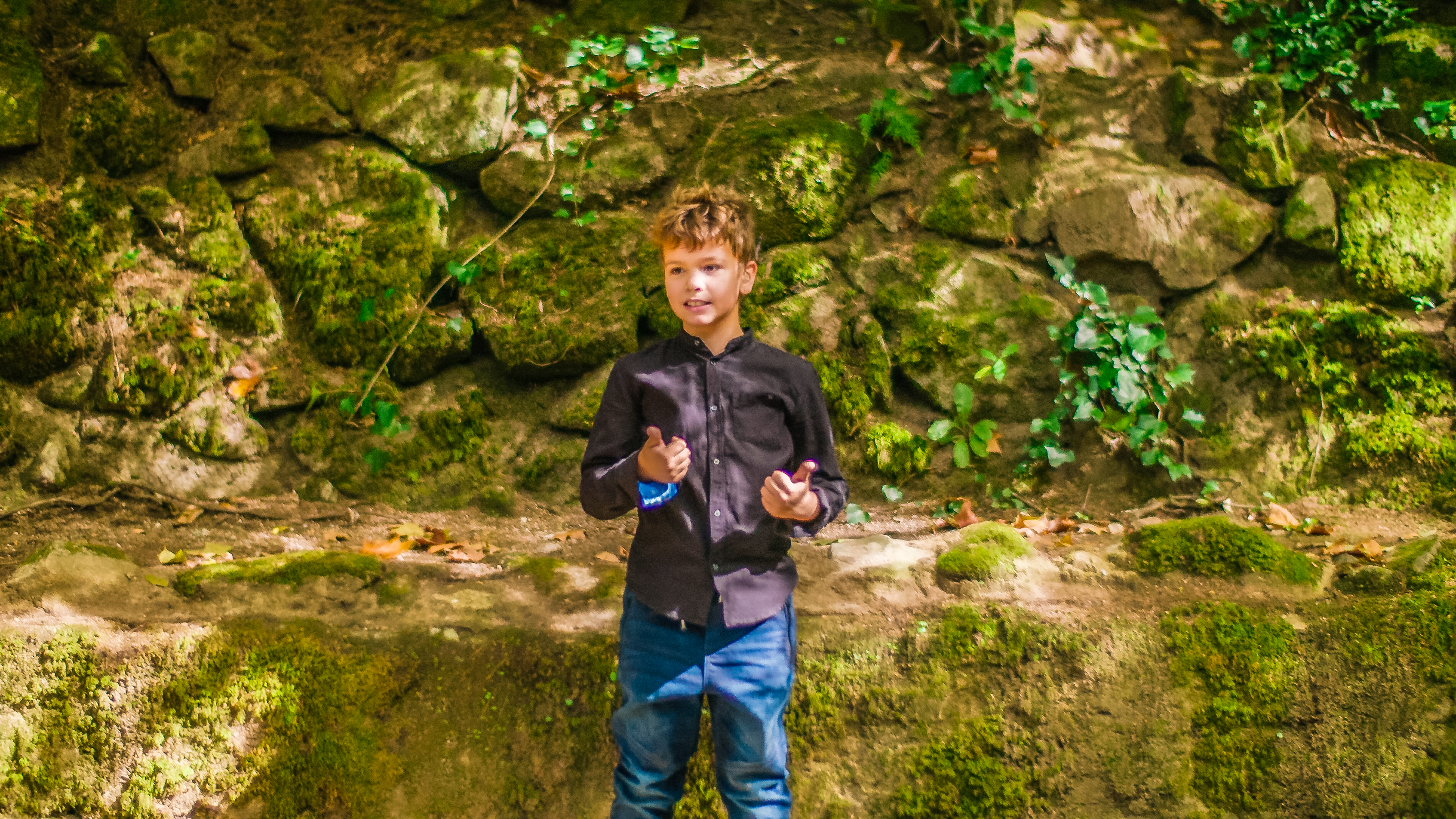 Tiago steht vor einer moosbewachsenen Felswand im Wald. Hier will er seine erste Unterrichtsstunde halten. Er trägt eine Jeans und ein dunkles Hemd und wirkt konzentriert und zuversichtlich.