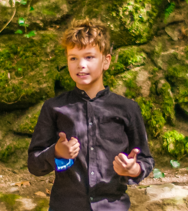 Tiago steht vor einer moosbewachsenen Felswand im Wald. Hier will er seine erste Unterrichtsstunde halten. Er trägt eine Jeans und ein dunkles Hemd und wirkt konzentriert und zuversichtlich.