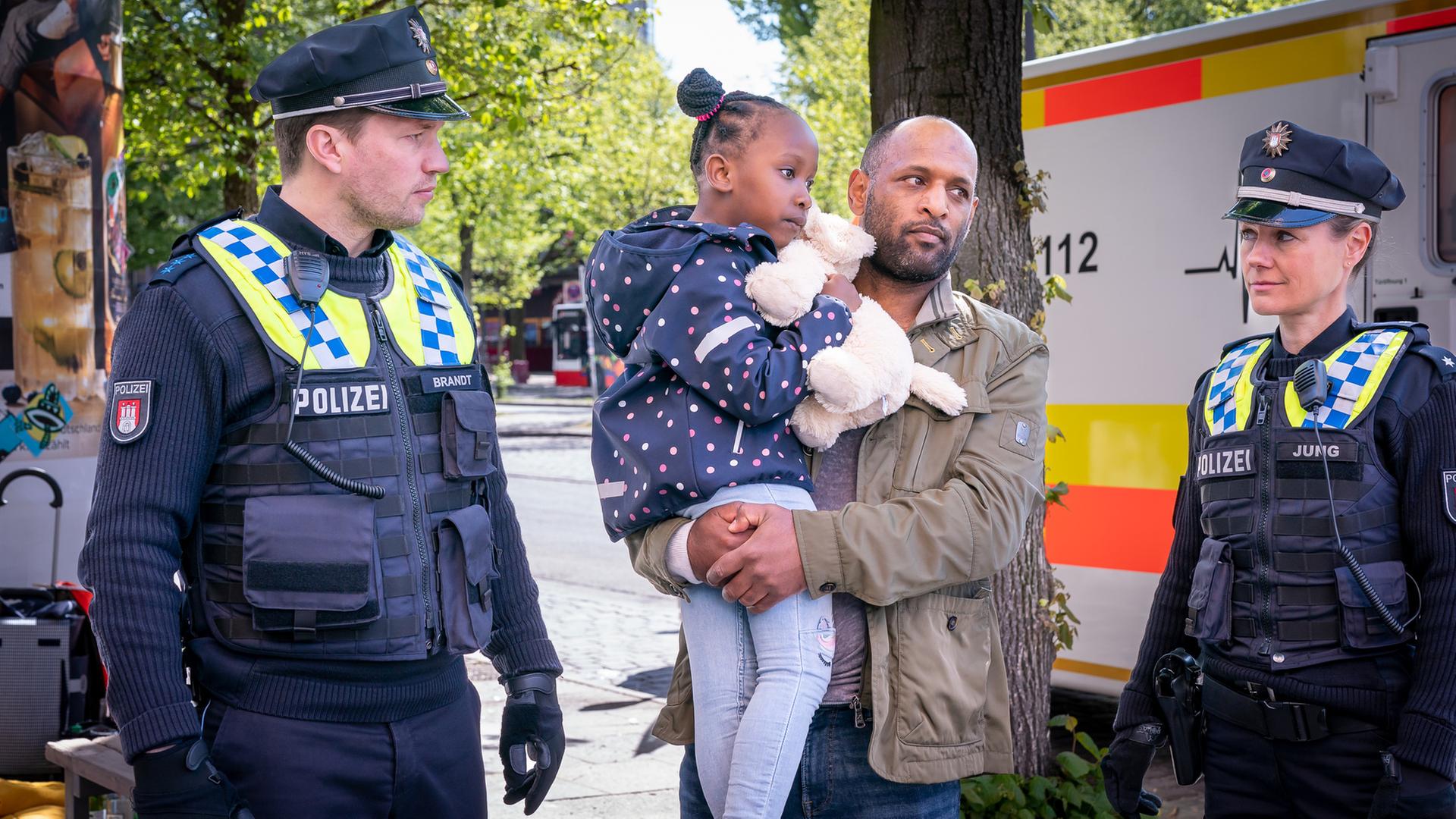 "Notruf Hafenkante - Tiefe Wunden": Kiano (Selam Tadese) steht zwischen der Polizistin Franzi Jung (Rhea Harder-Vennewald) und ihrem Kollegen Nick Brandt (Raúl Richter). Kiano hält seine Tochter Inaya (Sulaika Traore) in den Armen. Nick und Franzi schauen nachdenklich. Im Hintergrund ist ein Rettungswagen zu sehen.