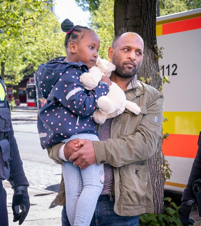 "Notruf Hafenkante - Tiefe Wunden": Kiano (Selam Tadese) steht zwischen der Polizistin Franzi Jung (Rhea Harder-Vennewald) und ihrem Kollegen Nick Brandt (Raúl Richter). Kiano hält seine Tochter Inaya (Sulaika Traore) in den Armen. Nick und Franzi schauen nachdenklich. Im Hintergrund ist ein Rettungswagen zu sehen.