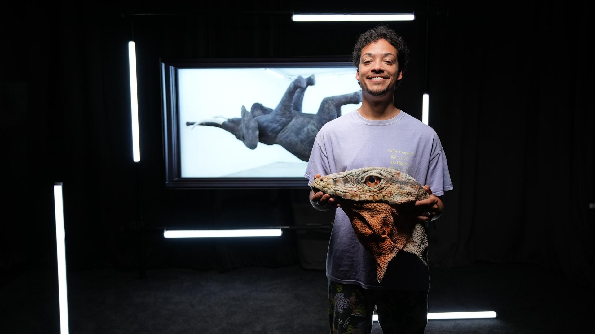 Ein Mann hält den Kopf einer überdimensionierten Echse in den Händen, er steht vor einem Foto eines Elefanten, der auf dem Rücken liegt oder schwimmt. 
