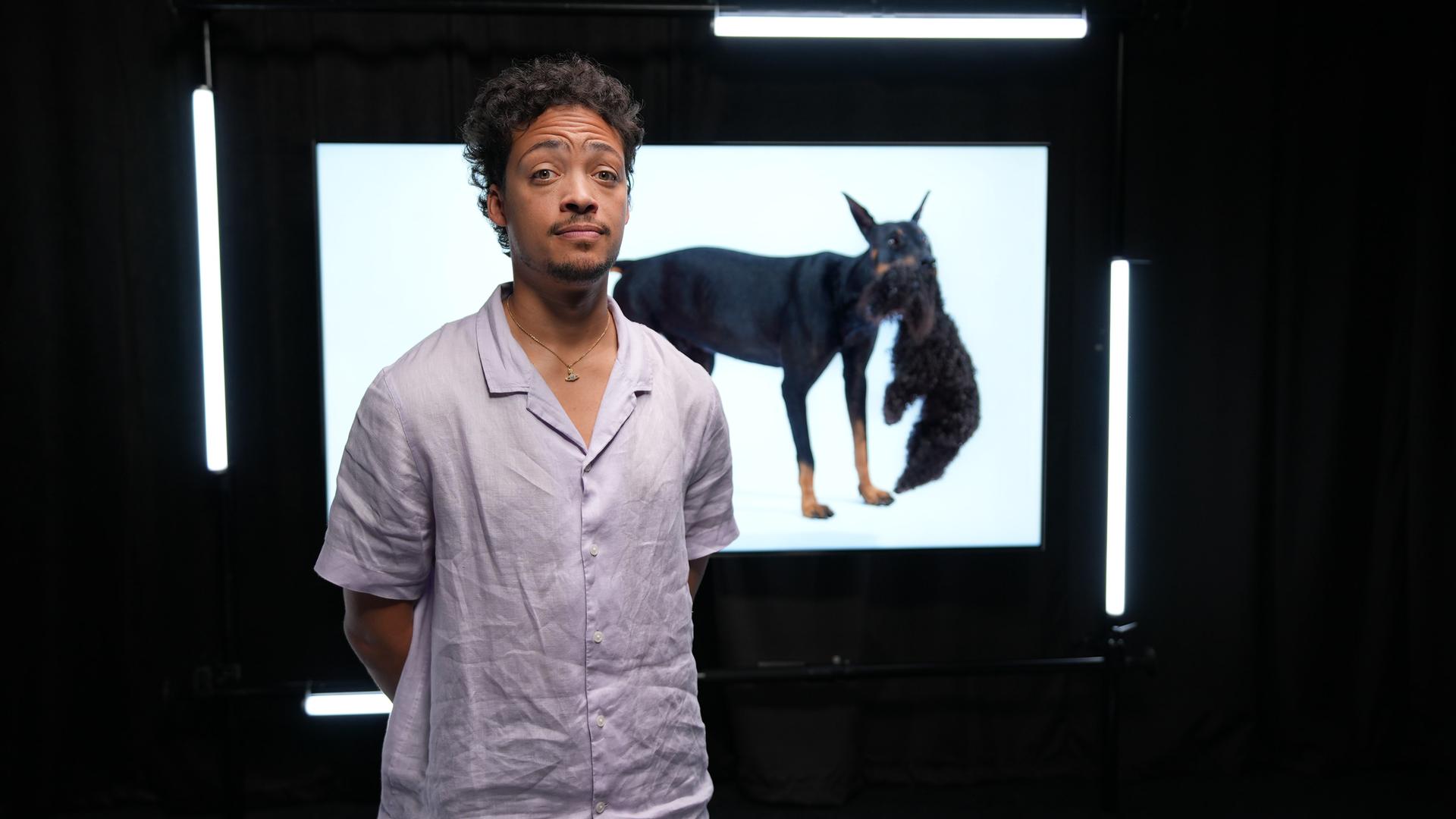 Ein Mann in hellem Hemd steht in einem dunklen Raum, hinter ihm sieht man ein Foto eines Dobermanns, der einen Pudel im Maul trägt. Der Mann legt die Stirn in Falten. 