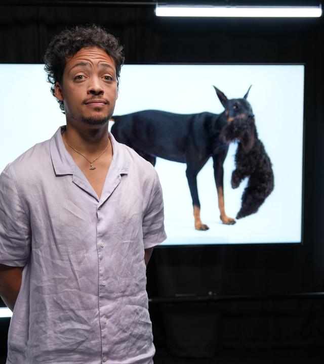 Ein Mann in hellem Hemd steht in einem dunklen Raum, hinter ihm sieht man ein Foto eines Dobermanns, der einen Pudel im Maul trägt. Der Mann legt die Stirn in Falten. 