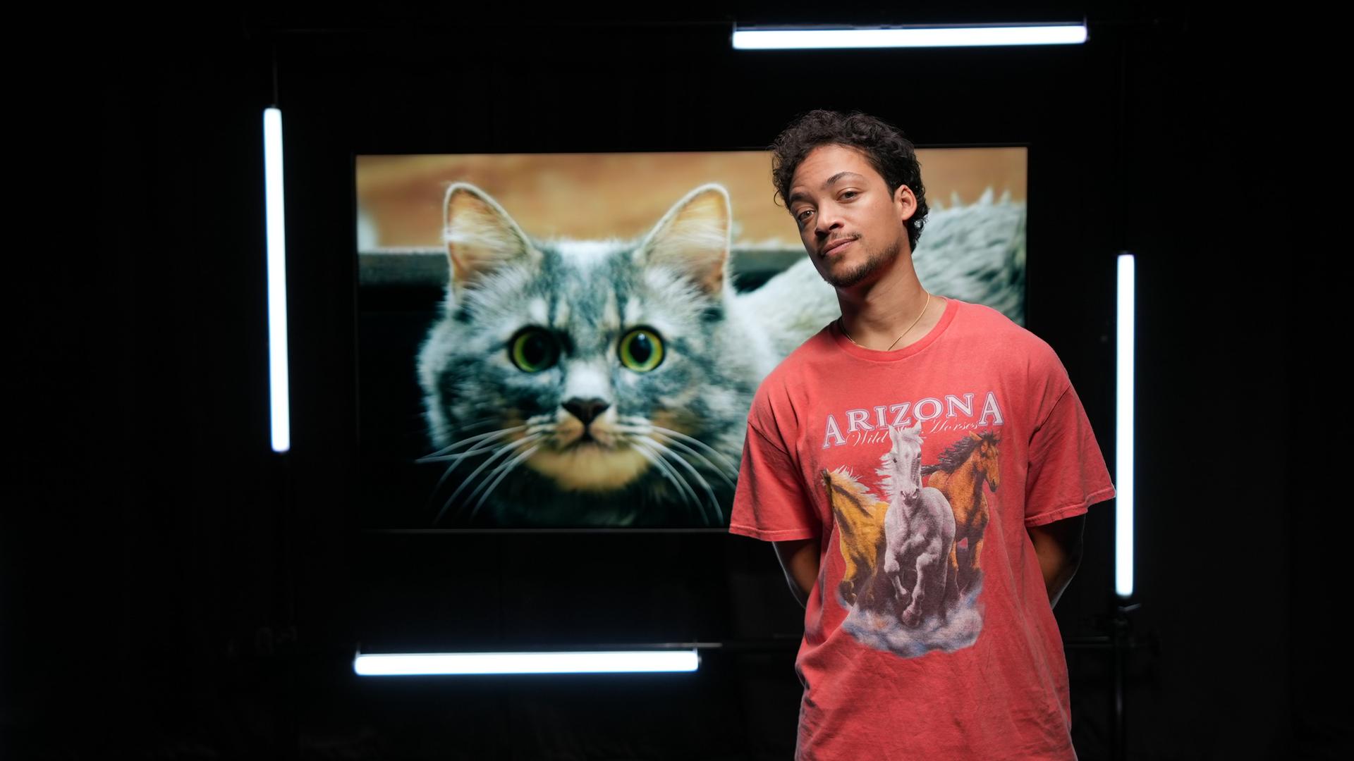 Ein Mann steht vor einem großen Foto einer Katze mit grünen Augen.  Er trägt ein rotes Shirt mit Pferden darauf.  