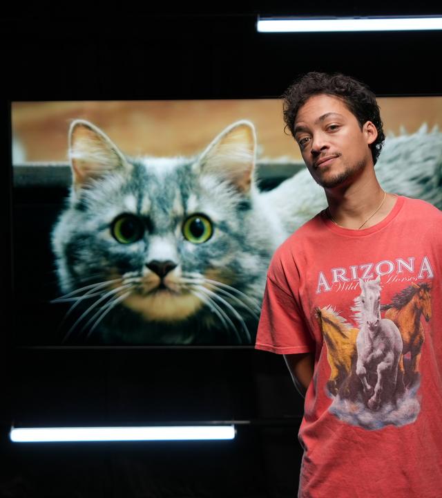 Ein Mann steht vor einem großen Foto einer Katze mit grünen Augen.  Er trägt ein rotes Shirt mit Pferden darauf.  