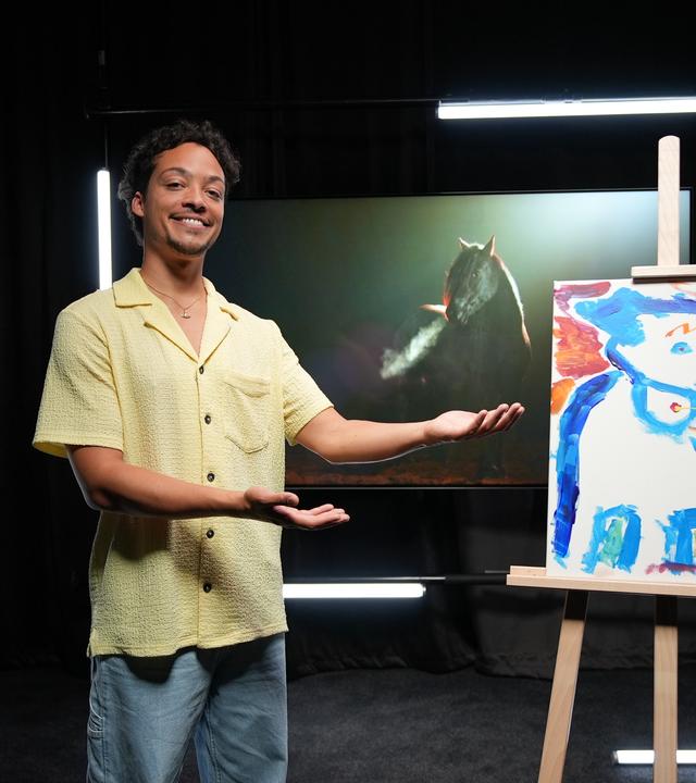 Ein Mann in gelbem Hemd präsentiert lächelnd ein Bild auf einer Staffelei. Drauf ist ein blaues Pferd zu sehen. Im Hintergrund sieht man ein großes Foto eines schwarzen Pferdes.