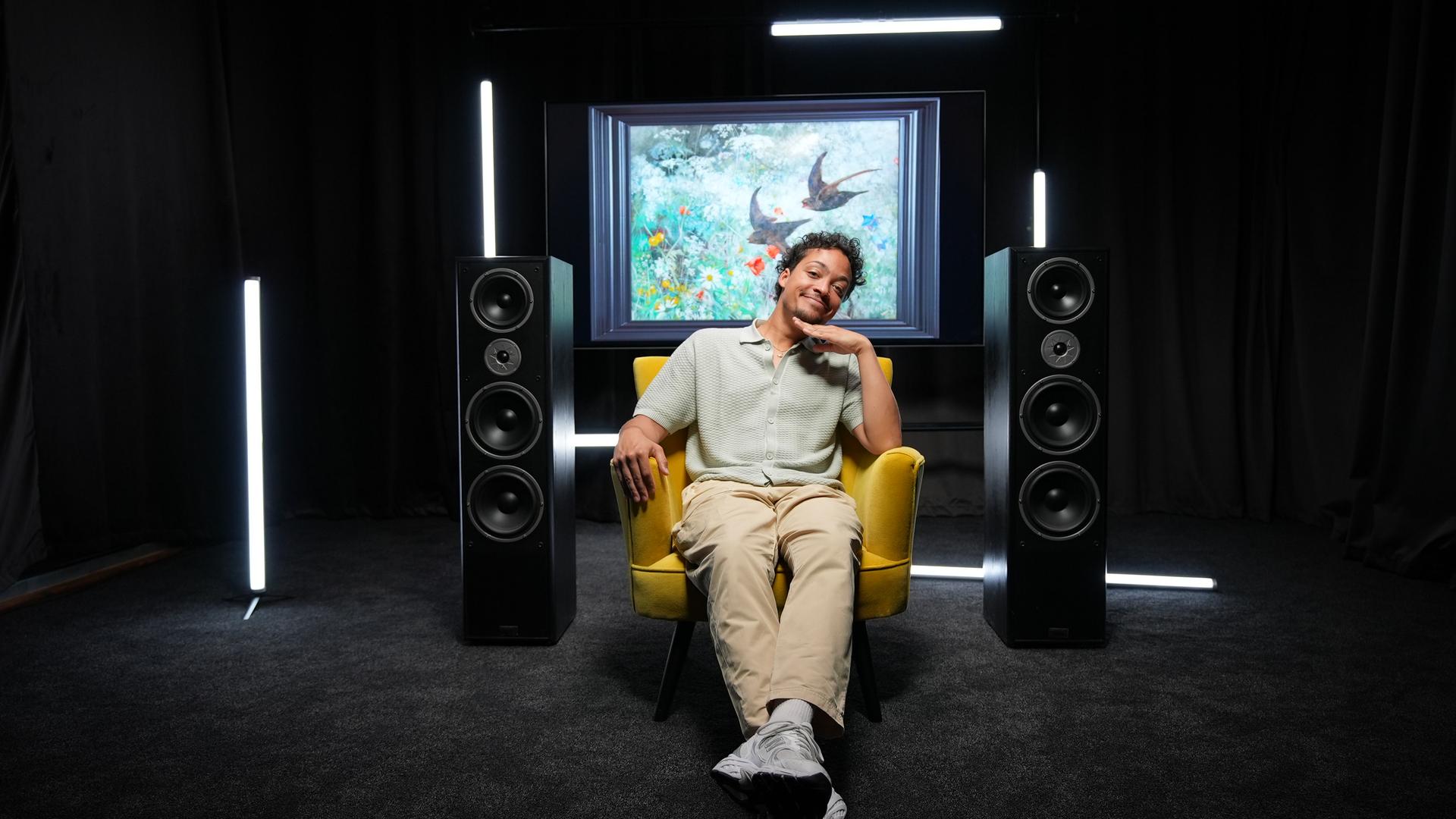 Ein Mann posiert in einem gelben Sessel in einem schwarzen Raum, links und rechts neben ihm stehen große Musik-Boxen, dahinter sieht man ein Kunstwerk mit Vögeln darauf.