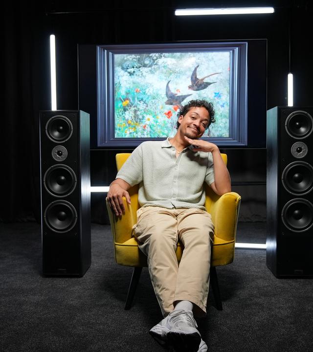 Ein Mann posiert in einem gelben Sessel in einem schwarzen Raum, links und rechts neben ihm stehen große Musik-Boxen, dahinter sieht man ein Kunstwerk mit Vögeln darauf.