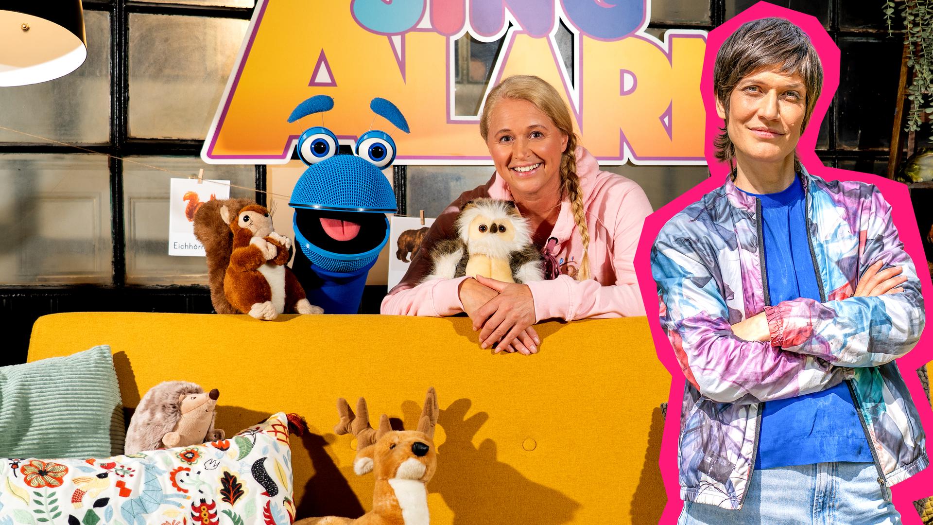 SingDing, Singa und Alin Coen sind vor dem SingAlarm Logo zu sehen. SingDing und Singa befinden sich hinter einer Couch, auf der zwei Kissen, ein Hirsch, ein Igel und ein Eichhörnchen-Stofftier platziert sind. Singa hält ein Eulen-Kuscheltier in den Armen.