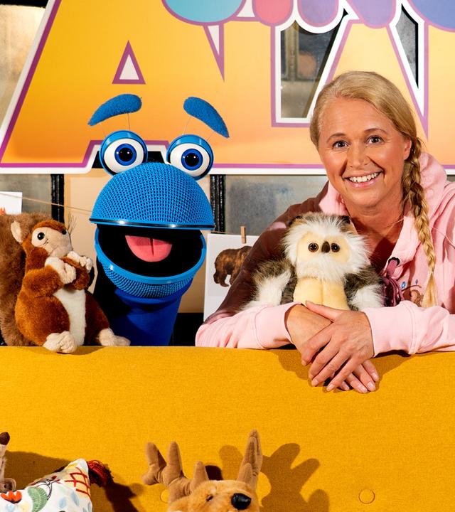 SingDing, Singa und Alin Coen sind vor dem SingAlarm Logo zu sehen. SingDing und Singa befinden sich hinter einer Couch, auf der zwei Kissen, ein Hirsch, ein Igel und ein Eichhörnchen-Stofftier platziert sind. Singa hält ein Eulen-Kuscheltier in den Armen.