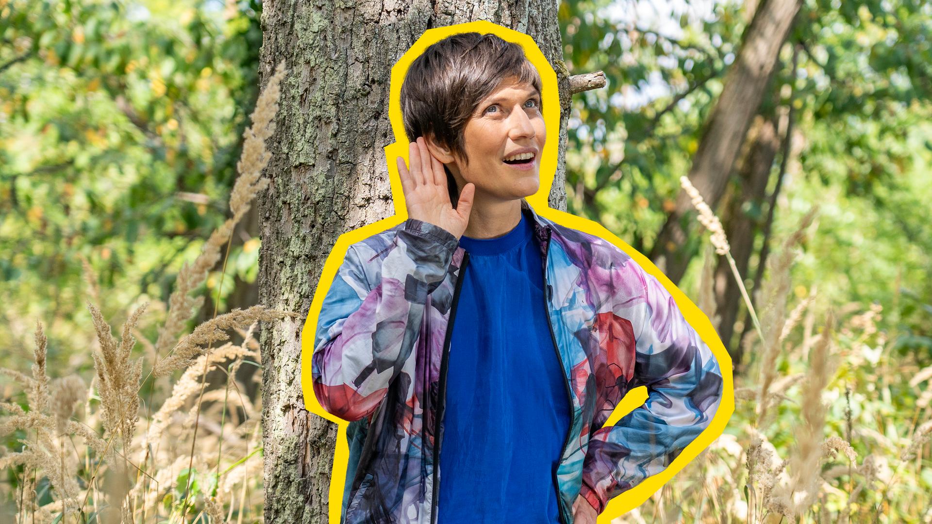 Alin Coen steht im Wald und hält sich ihre rechte Hand ans Ohr und lauscht den Naturgeräuschen.