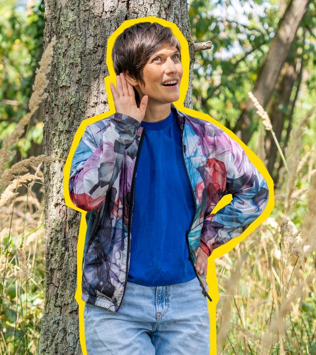 Alin Coen steht im Wald und hält sich ihre rechte Hand ans Ohr und lauscht den Naturgeräuschen.