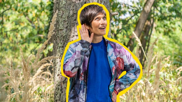 Alin Coen steht im Wald und hält sich ihre rechte Hand ans Ohr und lauscht den Naturgeräuschen.