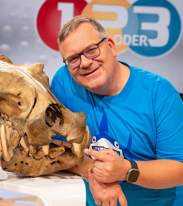 Dr. Markus Baur und Elton stehen an einem weißen Stehtisch. Auf dem Tisch steht zwischen ihnen ein großer Tierschädel. Beide zeigen auf die Zähne des Schädels.