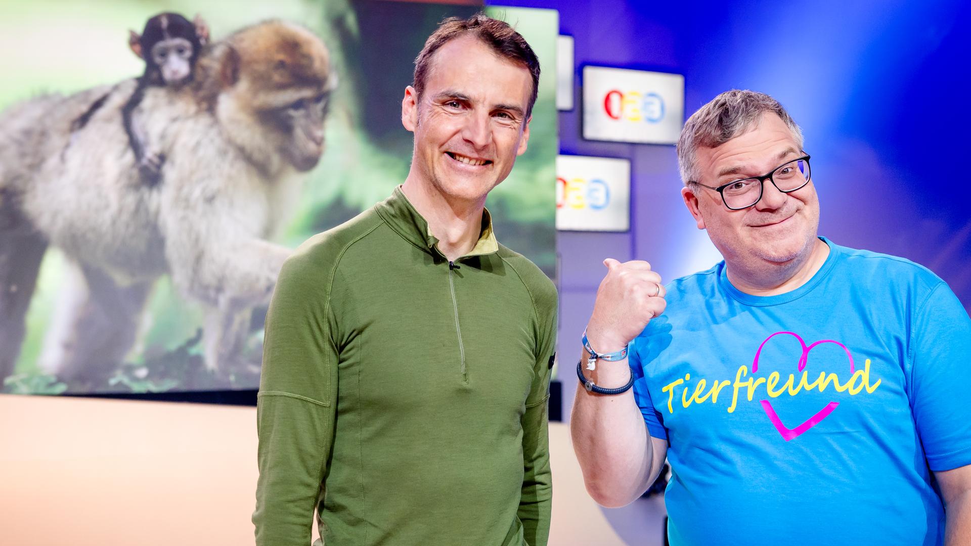 "1, 2 oder 3 - Tierisch toller Nachwuchs": Dr. Roland Hilgartner (l.) und Elton (r.) stehen im Studio vor dem Bild eines Berberaffen, der seinen Nachwuchs auf dem Rücken trägt.