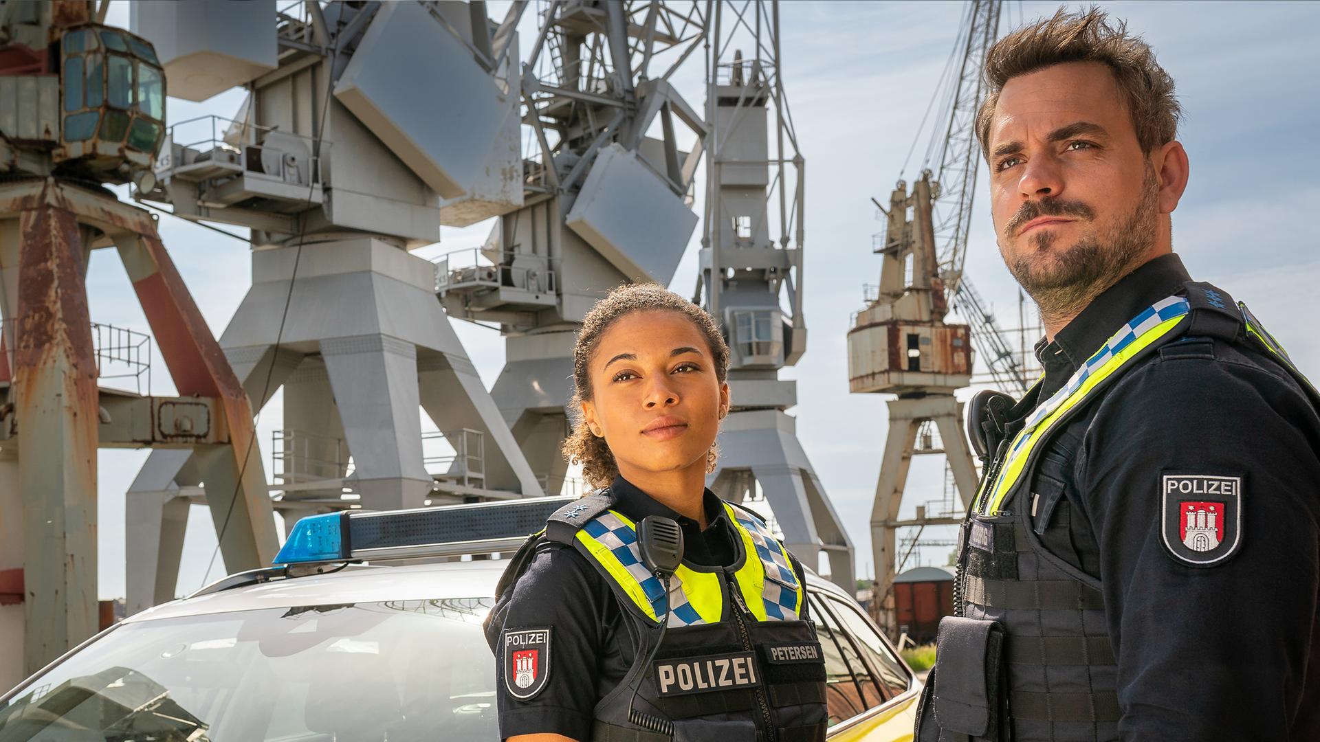 Die Polizistin Daisy Petersen (Aysha Joy Samuel) und ihr Kollege Kris Freiberg (Marc Barthel) stehen am Hafen neben ihrem Streifenwagen. Beide schauen in die gleiche Richtung.