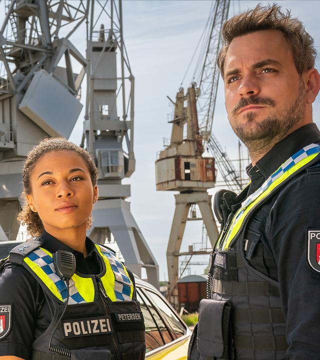 Die Polizistin Daisy Petersen (Aysha Joy Samuel) und ihr Kollege Kris Freiberg (Marc Barthel) stehen am Hafen neben ihrem Streifenwagen. Beide schauen in die gleiche Richtung.