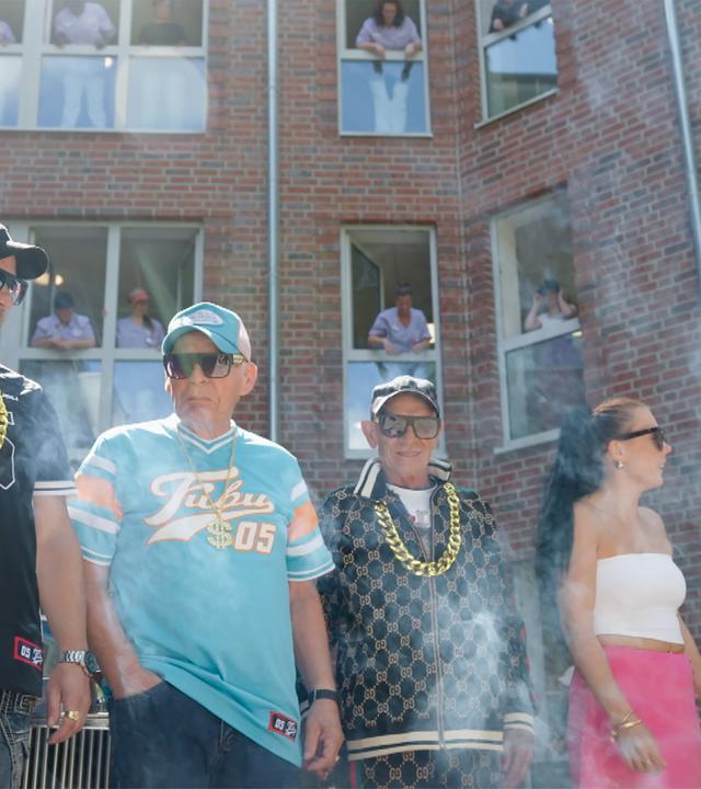 Drei wie Gangster-Rapper gekleidete Männer und zwei junge Frauen in engen Tops stehen vor einem Haus, in dessen Fenster jeweils eine Person steht und nach draußen schaut.