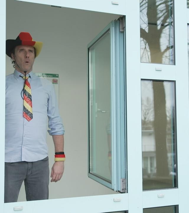 Reporter mit deutschen Accessoires (Hut, Krawatte) steht vor einem offenen Fenster.