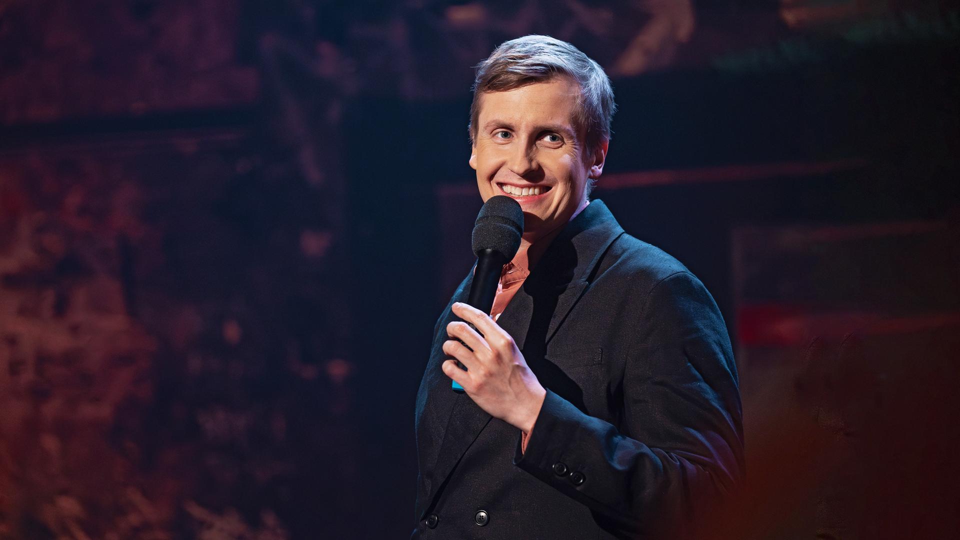 Till Reiners steht auf der rechten Seite des Bildes. Er lacht und hält in seiner linken Hand ein schwarzes Mikrofon. 