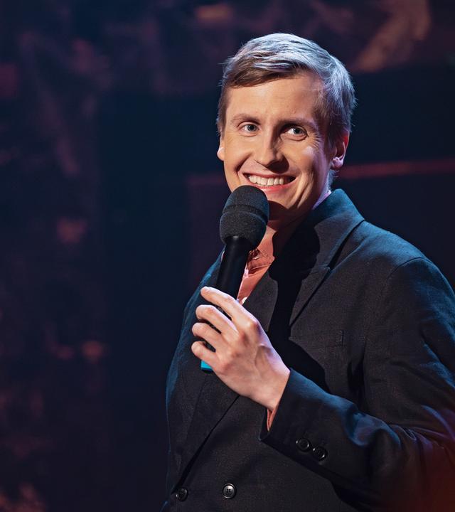 Till Reiners steht auf der rechten Seite des Bildes. Er lacht und hält in seiner linken Hand ein schwarzes Mikrofon. 