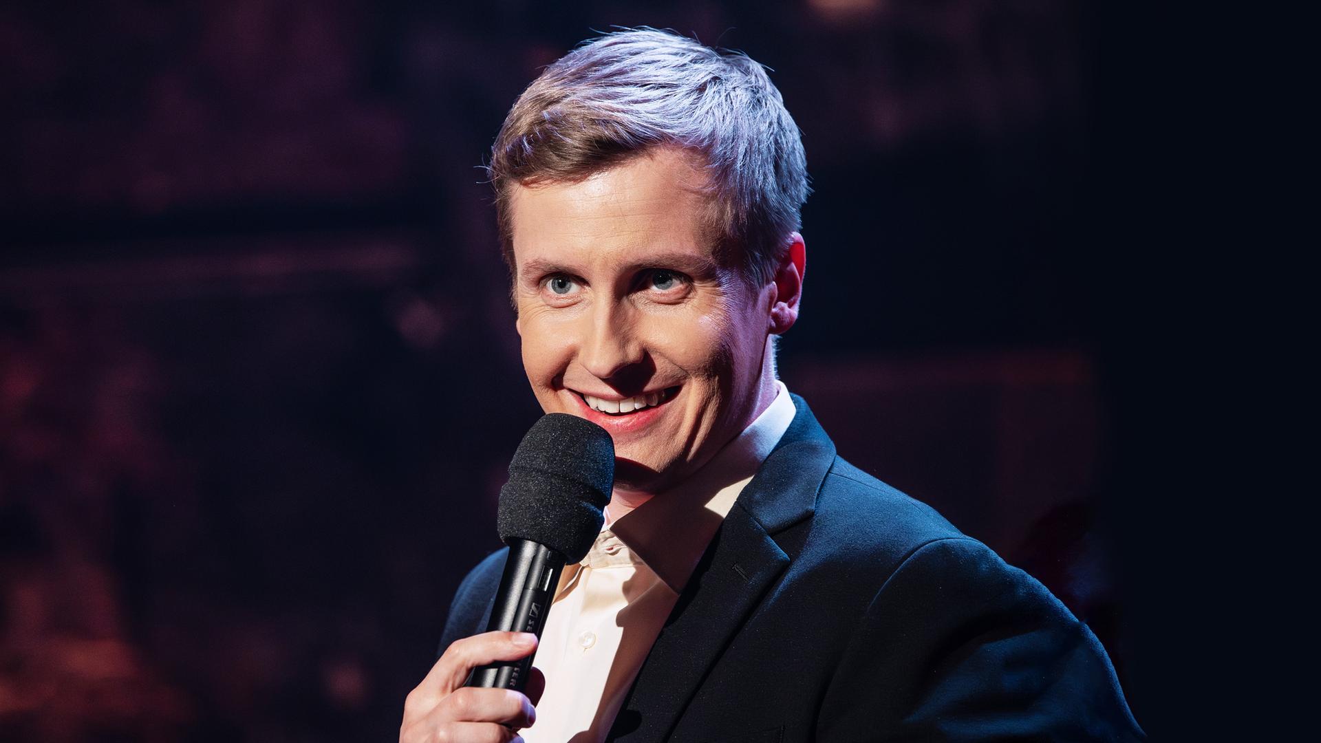 Till Reiners steht auf der rechten Seite des Bildes. Er lacht und hält in seiner linken Hand ein schwarzes Mikrofon.