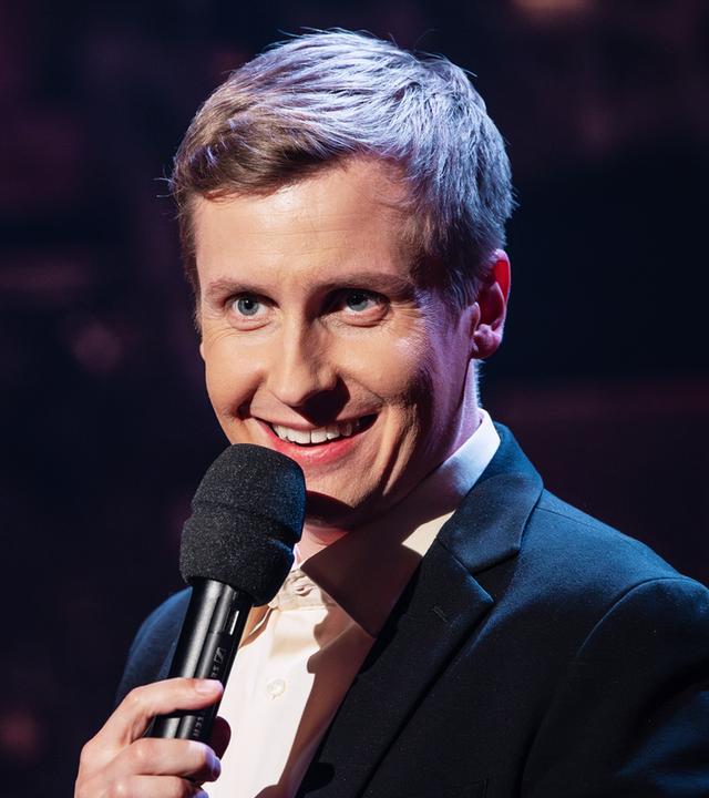 Till Reiners steht auf der rechten Seite des Bildes. Er lacht und hält in seiner linken Hand ein schwarzes Mikrofon.