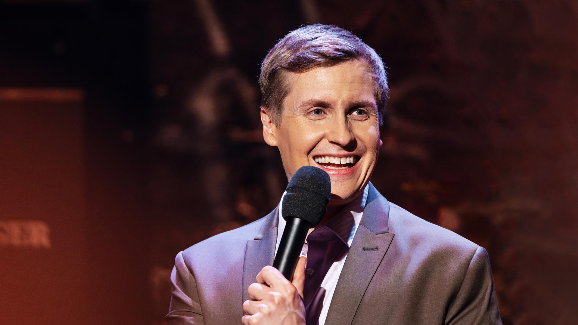 Till Reiners steht auf der rechten Seite des Bildes. Er lacht und hält in seiner linken Hand ein schwarzes Mikrofon