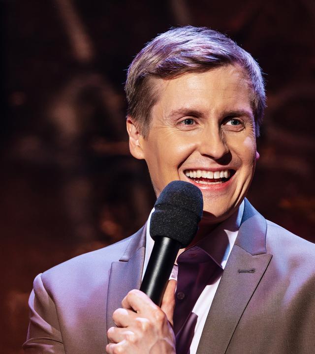 Till Reiners steht auf der rechten Seite des Bildes. Er lacht und hält in seiner linken Hand ein schwarzes Mikrofon