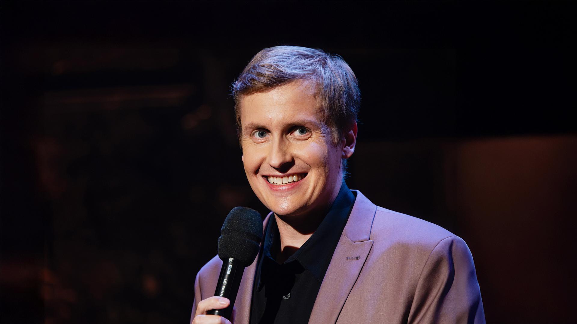 Till Reiners steht auf der rechten Seite des Bildes. Er lächelt und hält in seiner Hand ein schwarzes Mikrofon.