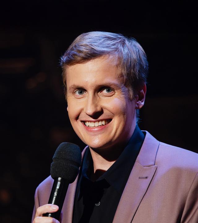 Till Reiners steht auf der rechten Seite des Bildes. Er lächelt und hält in seiner Hand ein schwarzes Mikrofon.