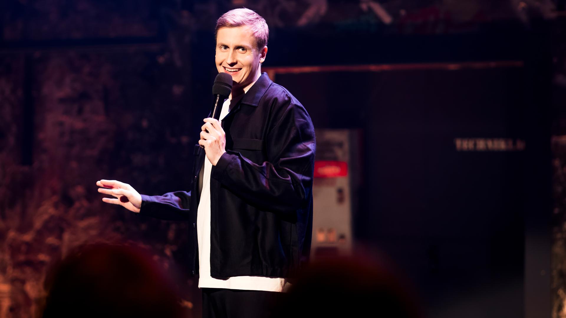 Till Reiners ist auf der linken Bildseite zu sehen. Till Reiners lächelt und schaut in Richtung der rechten Bildseite. In seiner linken Hand hält er ein Mikrofon. 