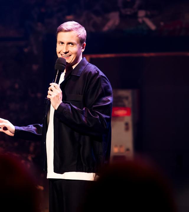 Till Reiners ist auf der linken Bildseite zu sehen. Till Reiners lächelt und schaut in Richtung der rechten Bildseite. In seiner linken Hand hält er ein Mikrofon. 