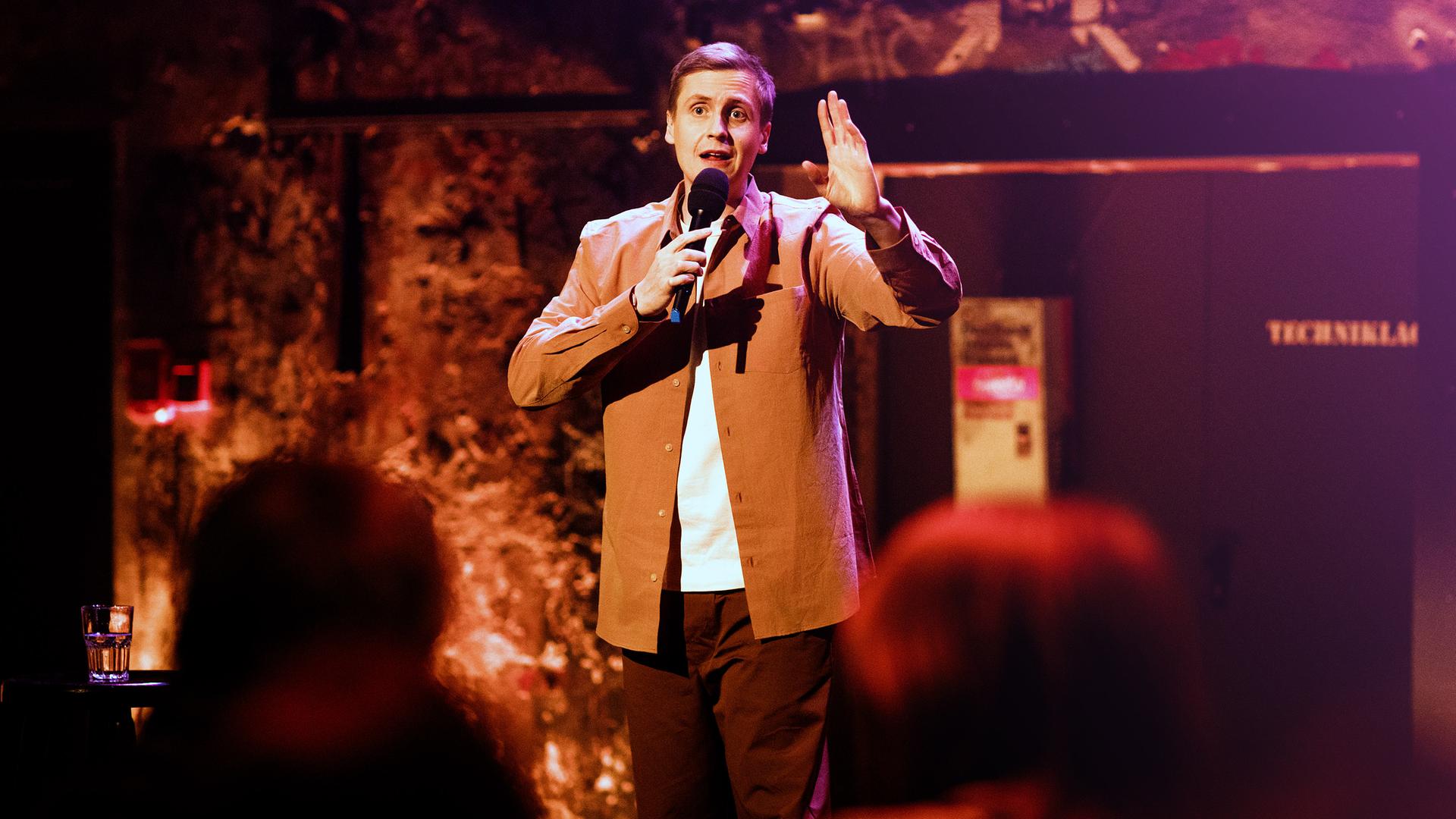 Till Reiners in der Mitte des Bildes. In seiner rechten Hand hält er ein Mikrofon. Die Beleuchtung ist rot-orange.