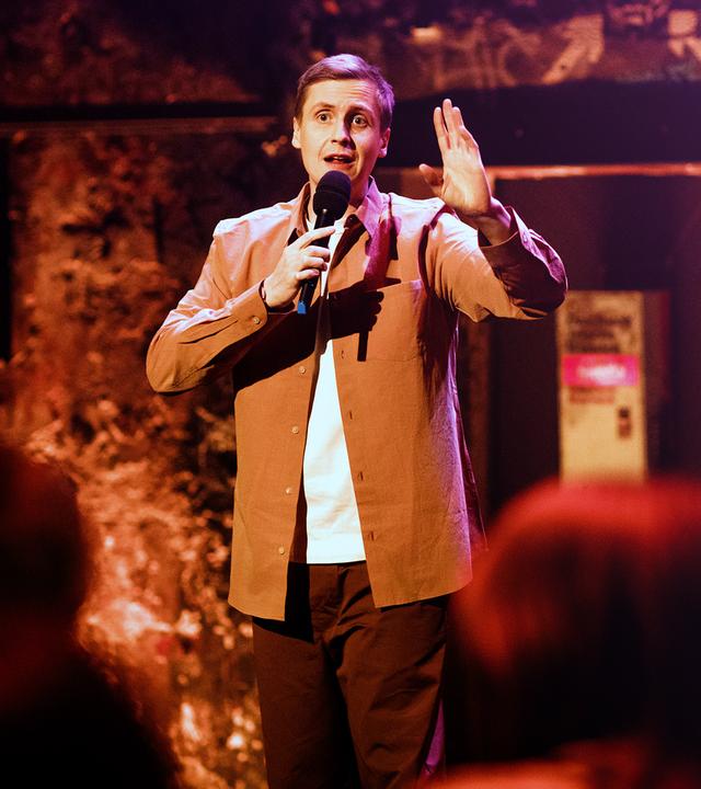 Till Reiners in der Mitte des Bildes. In seiner rechten Hand hält er ein Mikrofon. Die Beleuchtung ist rot-orange.