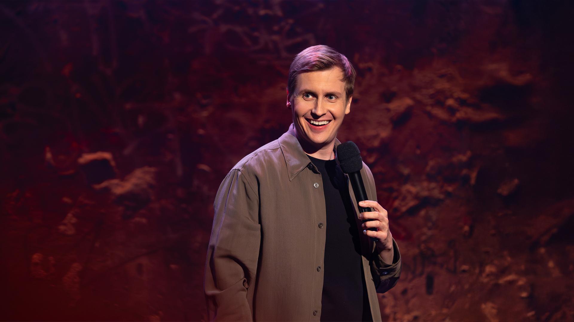 Till Reiners ist auf der rechten Bildseite zu sehen. Till Reiners lächelt und schaut in Richtung der linken Bildseite. In seiner linken Hand hält er ein Mikrofon. Die Beleuchtung ist rot und lila.