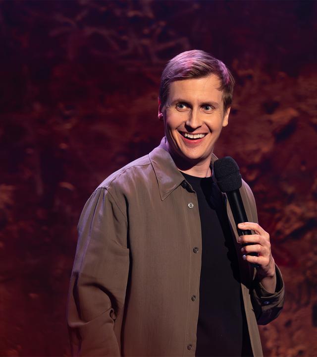 Till Reiners ist auf der rechten Bildseite zu sehen. Till Reiners lächelt und schaut in Richtung der linken Bildseite. In seiner linken Hand hält er ein Mikrofon. Die Beleuchtung ist rot und lila.