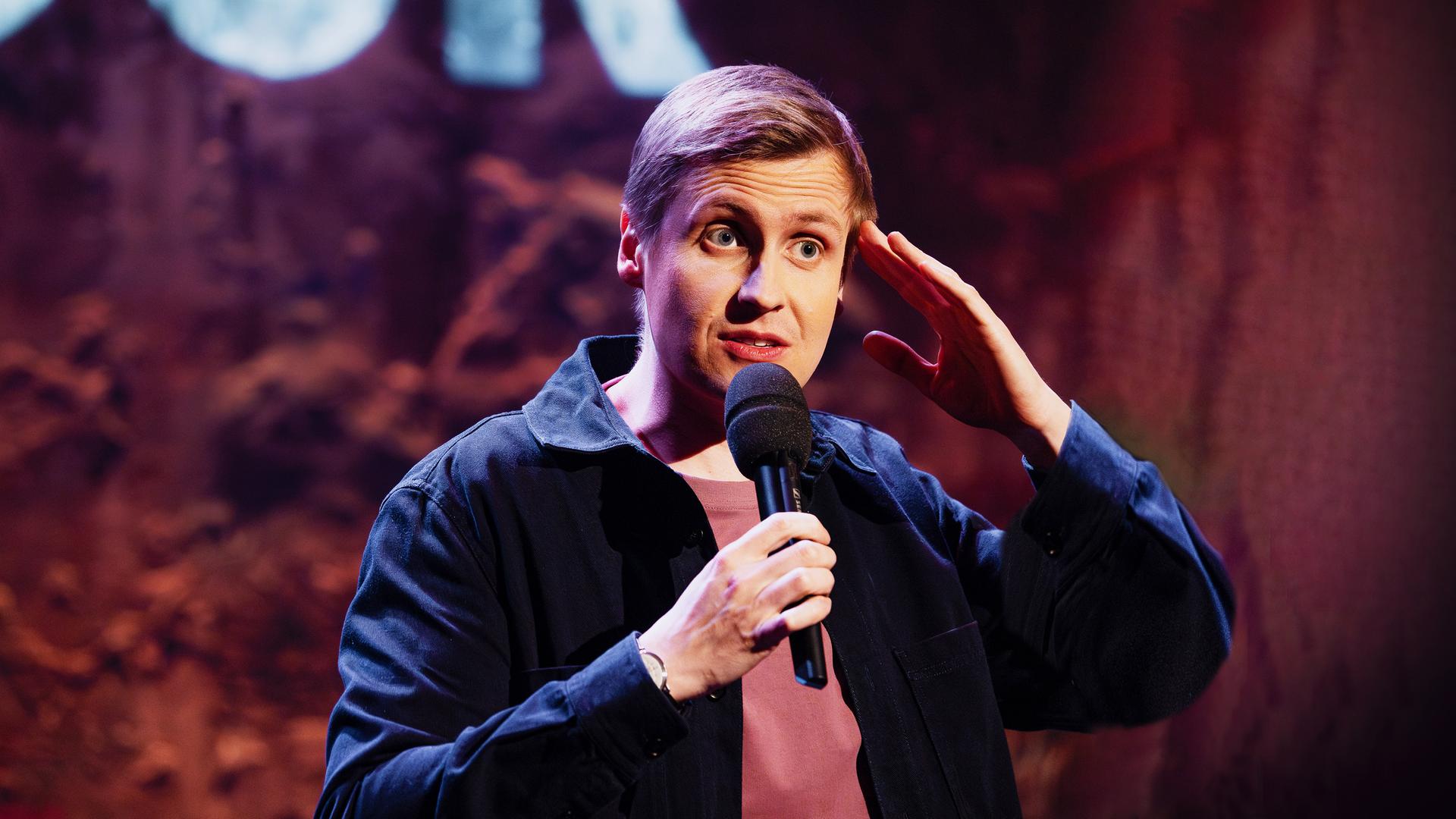 Till Reiners ist auf der rechten Bildseite zu sehen. Er greift sich mit seiner linken Hand an die Stirn. In der rechten Hand hält er sein Mikrofon.