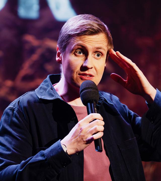 Till Reiners ist auf der rechten Bildseite zu sehen. Er greift sich mit seiner linken Hand an die Stirn. In der rechten Hand hält er sein Mikrofon.