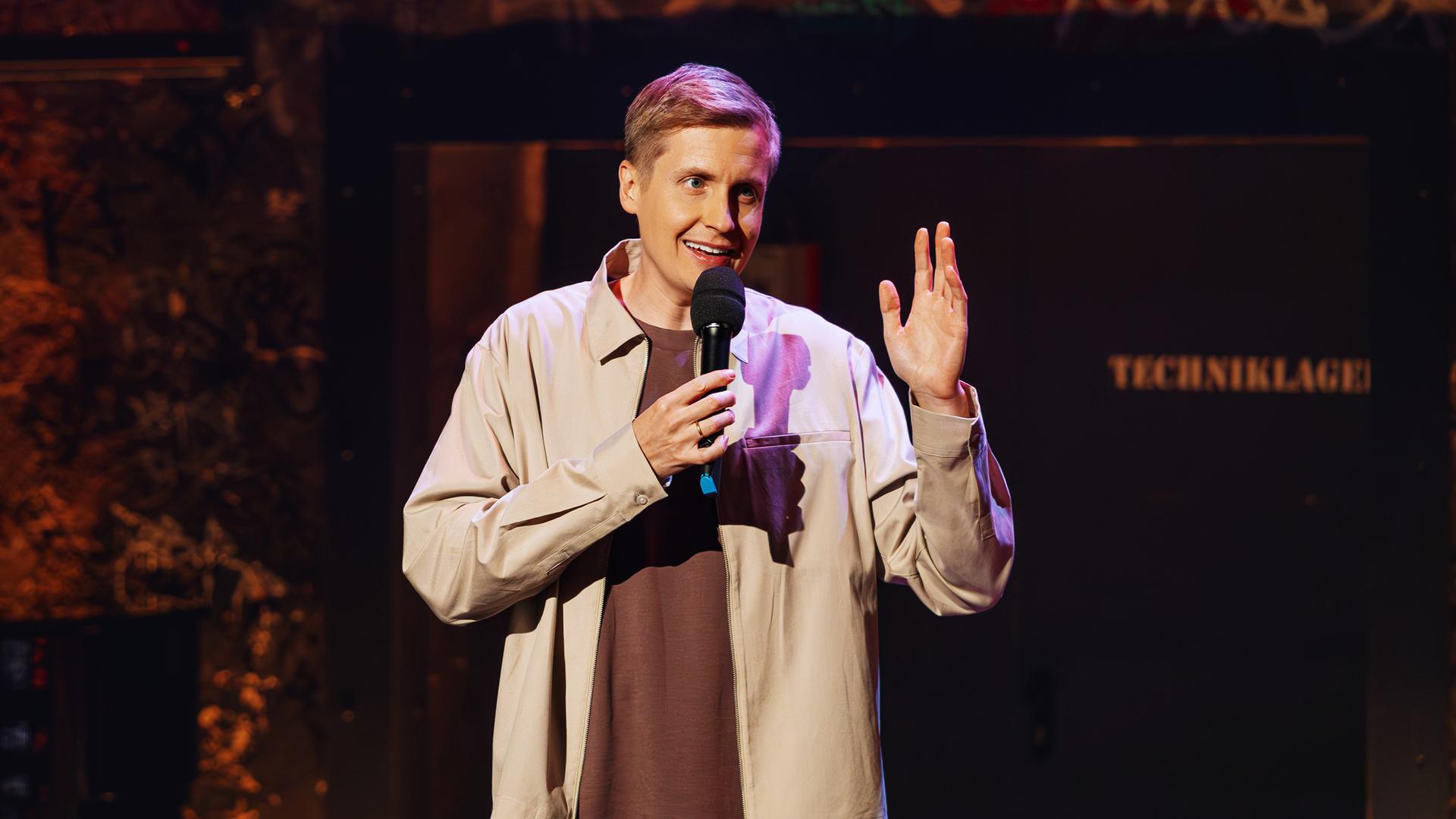 Till Reiners hält ein Mikrofon in einer Hand, mit der anderen Hand gestikuliert er. Er trägt ein braunes Shirt und darüber eine beige Jacke.