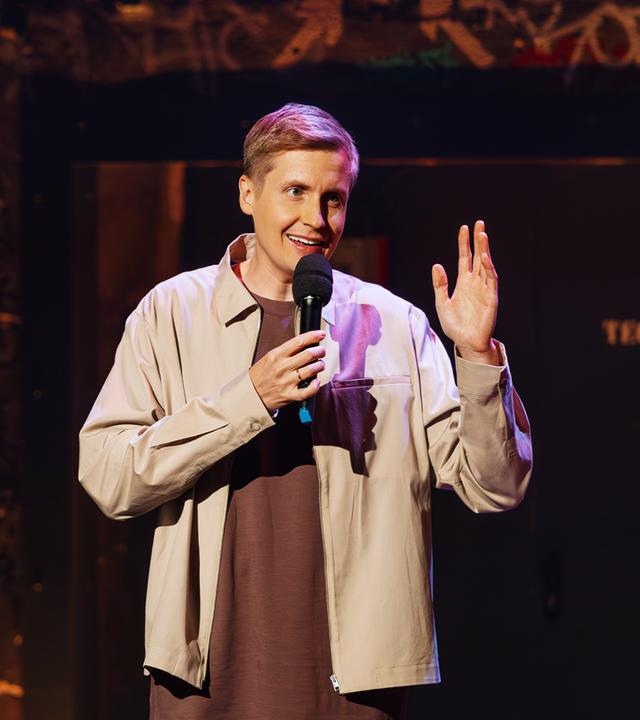 Till Reiners hält ein Mikrofon in einer Hand, mit der anderen Hand gestikuliert er. Er trägt ein braunes Shirt und darüber eine beige Jacke.